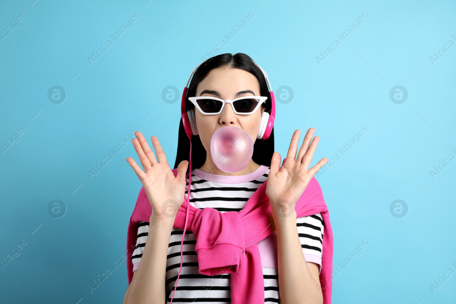 Photo of Fashionable young woman with headphones blowing bubblegum on light blue background