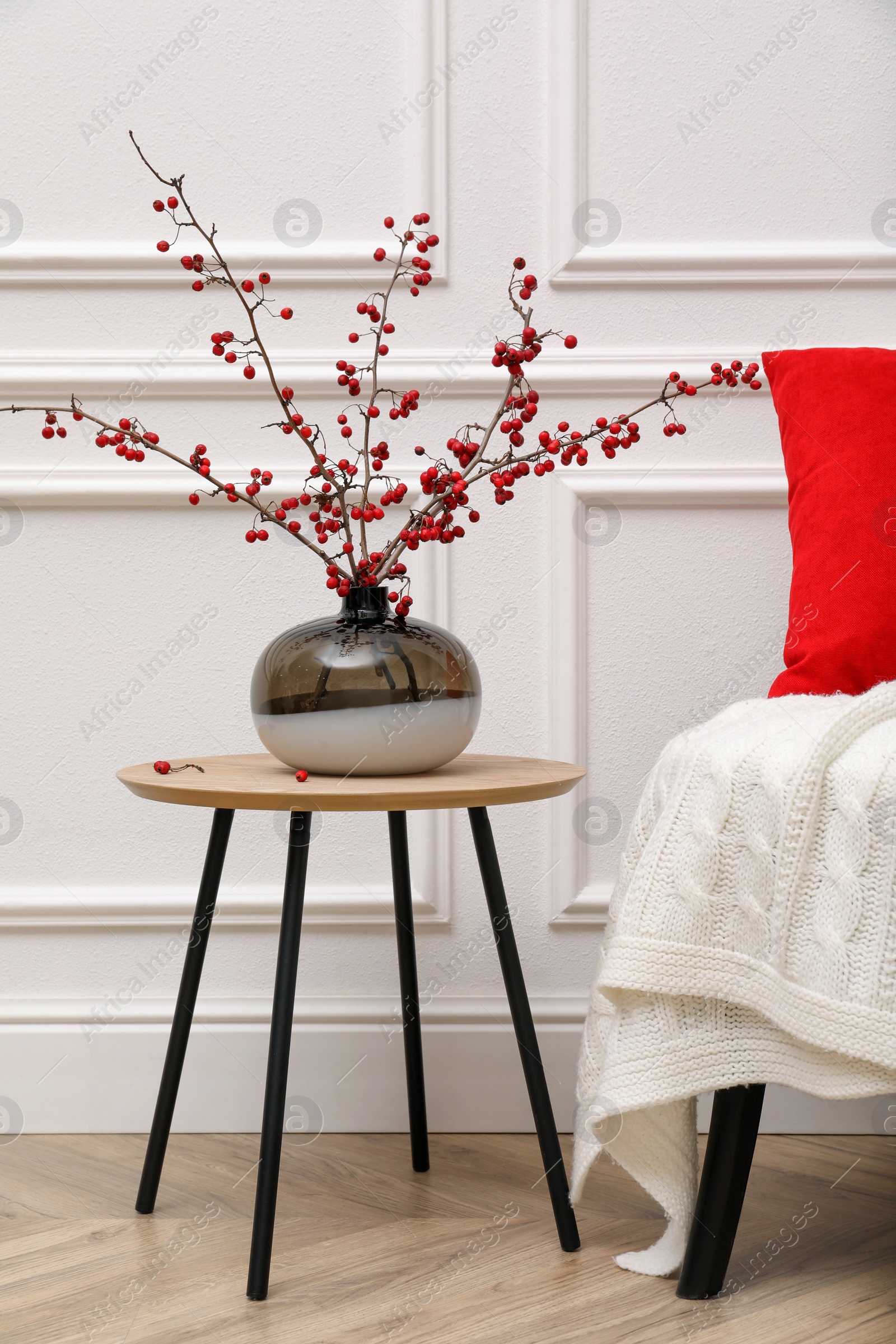 Photo of Hawthorn branches with red berries in vase on table near white wall indoors