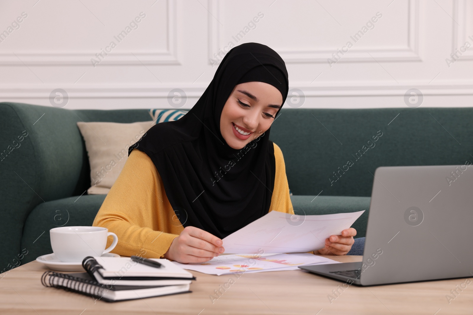 Photo of Muslim woman in hijab writing notes near laptop at wooden table indoors