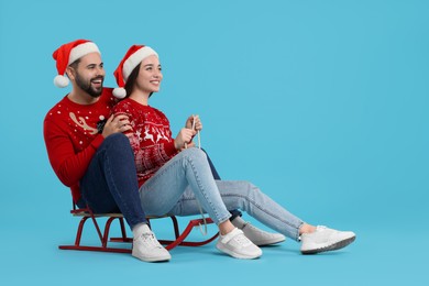 Happy young couple in Christmas sweaters and Santa hats on sled against light blue background. Space for text