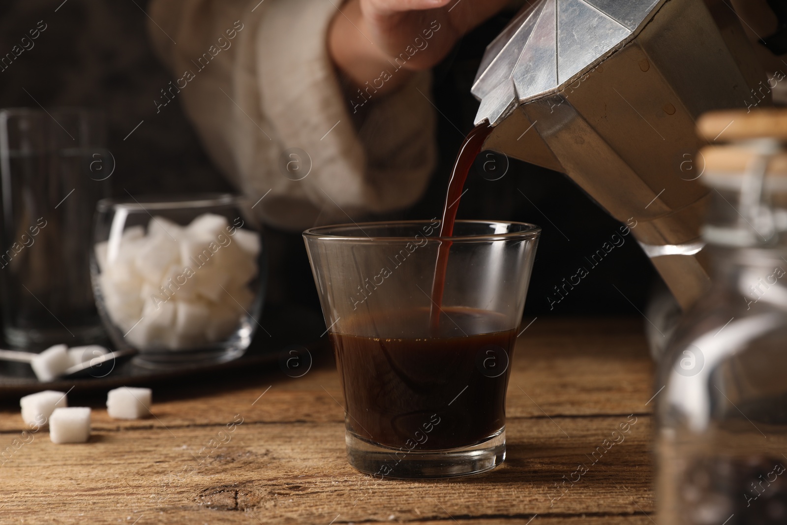 Photo of Woman pouring aromatic coffee from moka pot into glass at wooden table, closeup