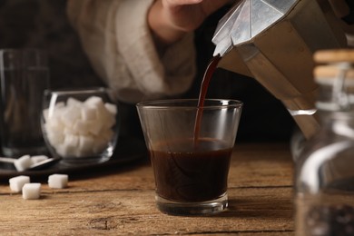 Woman pouring aromatic coffee from moka pot into glass at wooden table, closeup