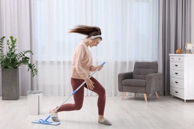 Photo of Enjoying cleaning. Happy woman in headphones dancing with mop indoors