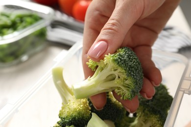 Woman putting broccoli into container at table, closeup. Food storage