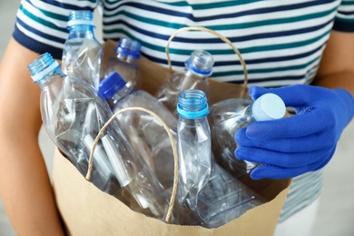 Photo of Woman holding paper bag with used plastic bottles indoors, closeup. Recycling problem