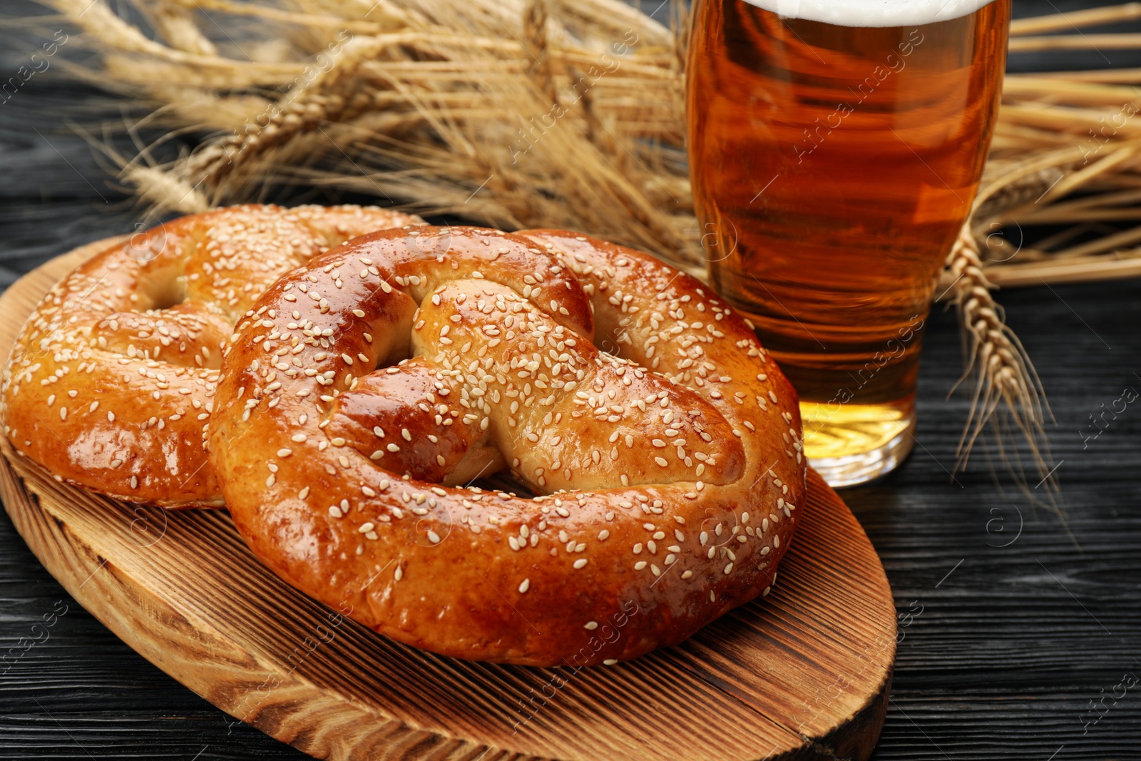 Photo of Tasty pretzels, glass of beer and wheat spikes on black wooden table