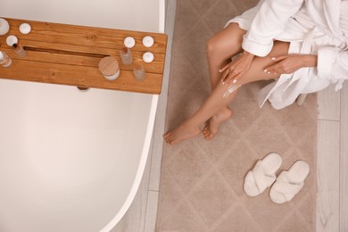 Woman applying body cream onto her smooth legs in bathroom, above view