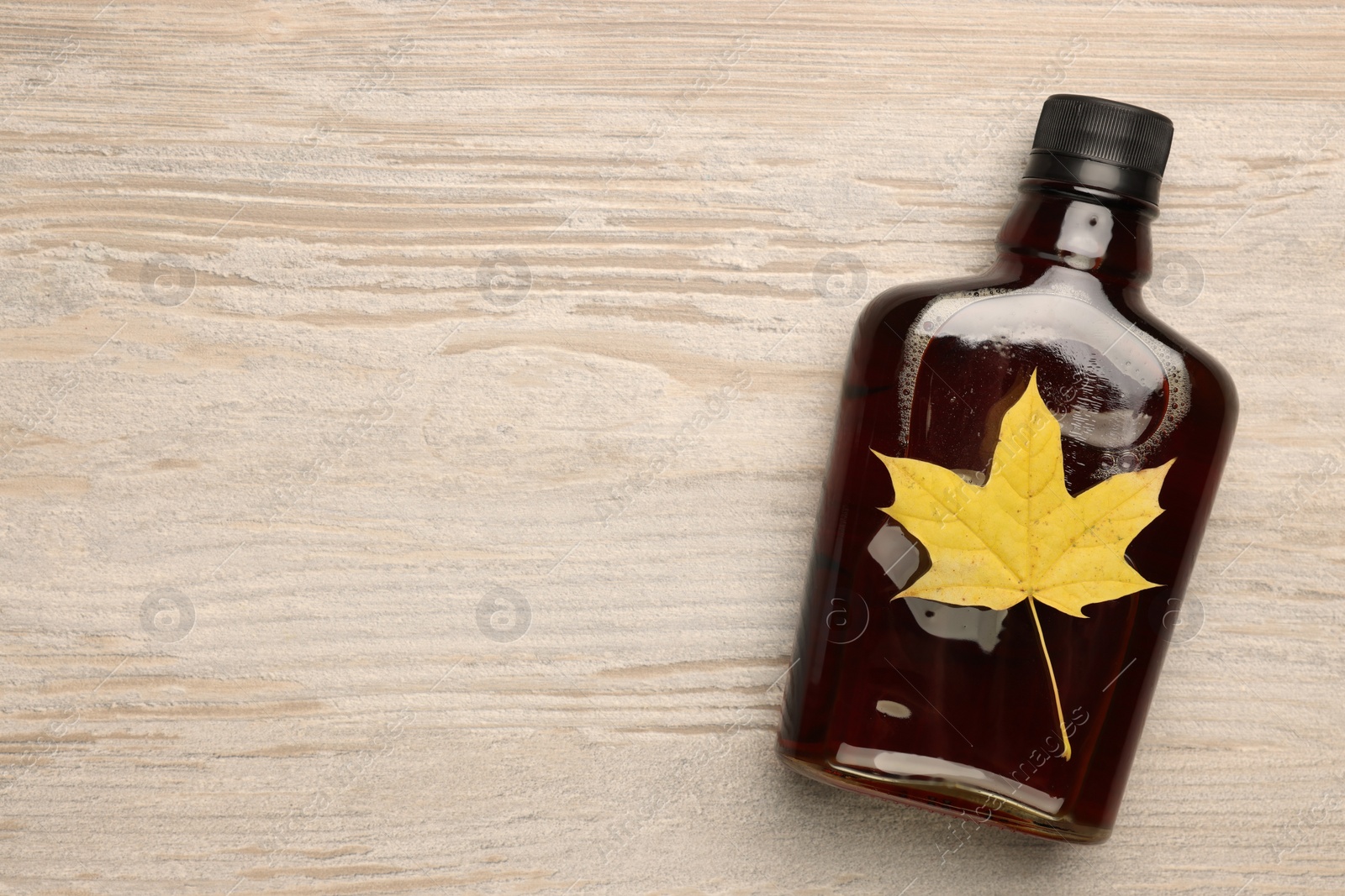 Photo of Glass bottle of tasty maple syrup and dry leaf on wooden table, top view. Space for text