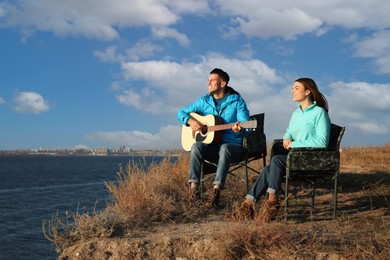 Couple with guitar resting in camping chairs near river on sunny day