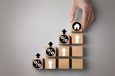 Image of Mortgage rate rising illustrated by upward arrows. Woman putting wooden cube with house icon near other ones on light grey background, top view