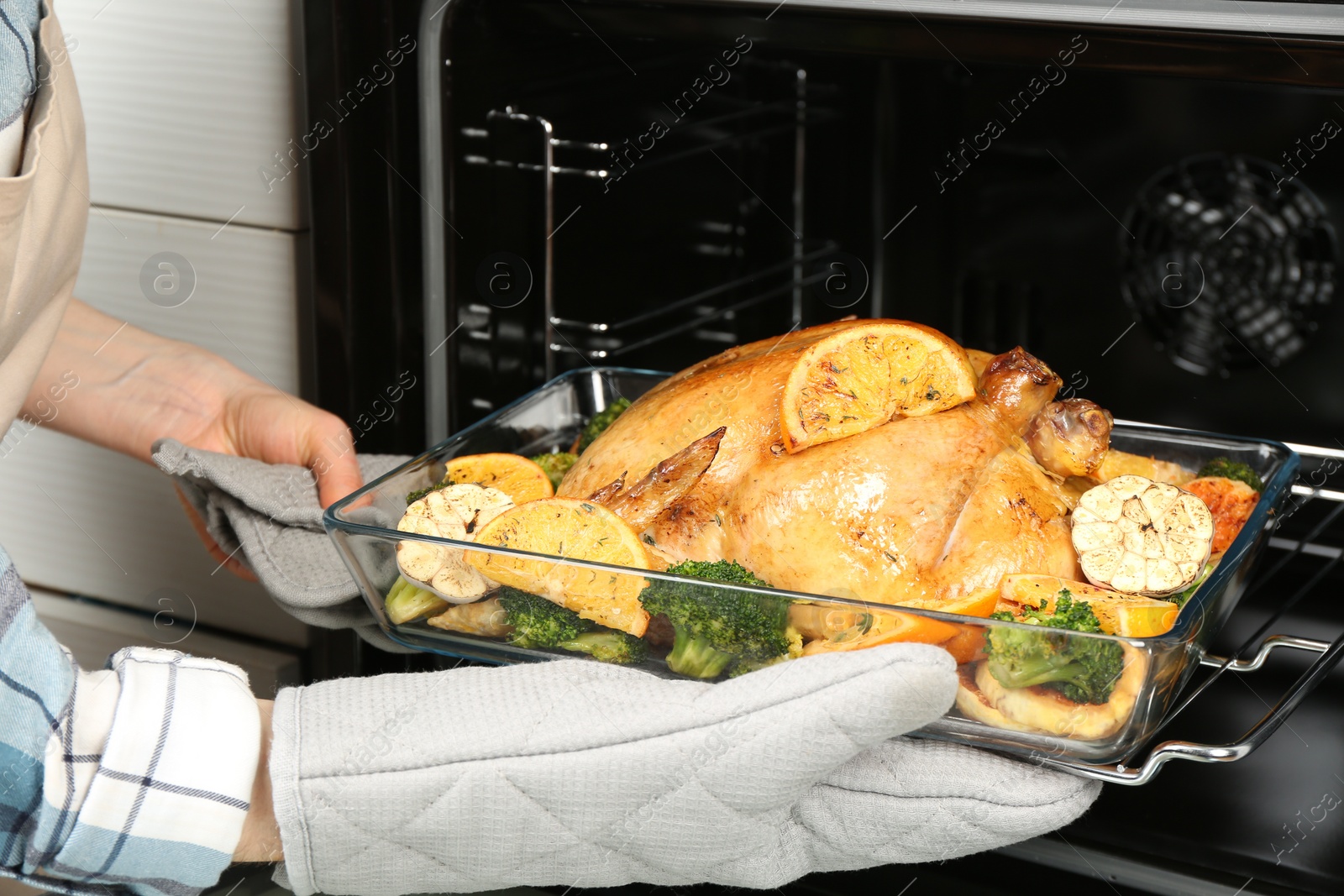 Photo of Woman taking baked chicken with oranges and vegetables out of oven, closeup