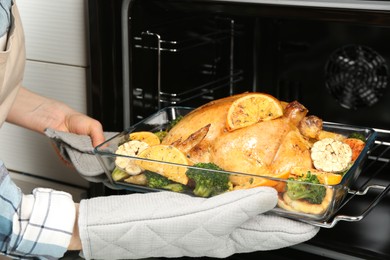 Photo of Woman taking baked chicken with oranges and vegetables out of oven, closeup