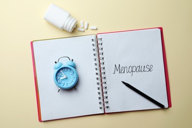 Photo of Notebook with word Menopause, alarm clock, pills and marker on beige background, flat lay