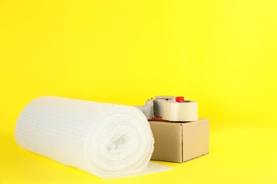 Photo of Bubble wrap roll, tape dispenser and cardboard box on yellow background, space for text