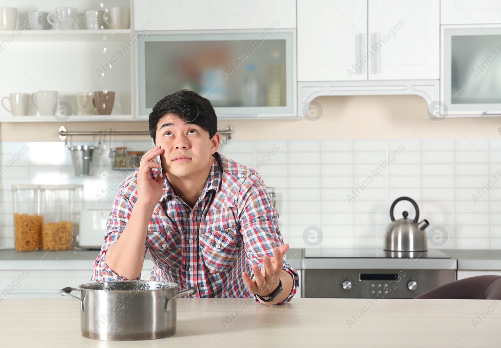 Photo of Emotional man calling plumber near table with saucepan under leaking water from ceiling in kitchen