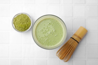 Photo of Glass of tasty matcha smoothie, powder and bamboo whisk on white tiled table, flat lay