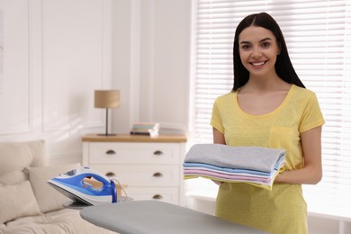 Photo of Woman with folded clothes near ironing board at home. Space for text