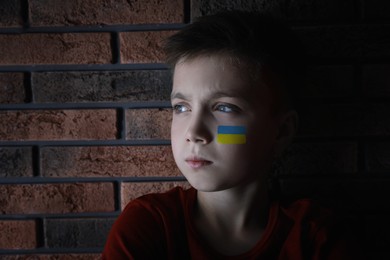 Image of Sad little boy with picture of Ukrainian flag on cheek near brick wall. Stop war in Ukraine