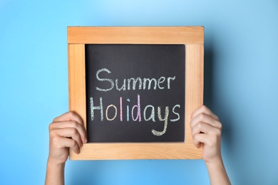 Photo of Schoolgirl holding blackboard with text SUMMER HOLIDAYS on light blue background, closeup
