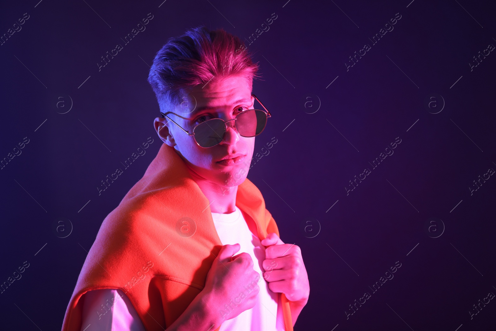 Photo of Stylish young man in sunglasses on dark background in neon lights. Space for text