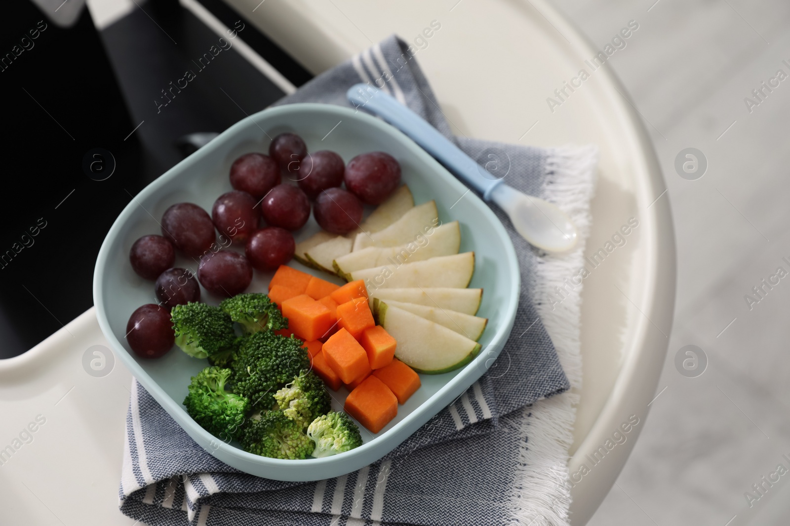 Photo of High chair with food in baby tableware on tray indoors, closeup. Above view