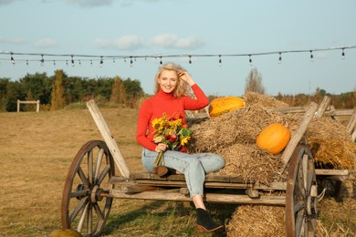 Beautiful woman with bouquet sitting on wooden cart with pumpkins and hay in field. Autumn season