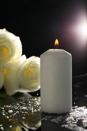 White roses and burning candle on black mirror surface in darkness, closeup. Funeral symbols
