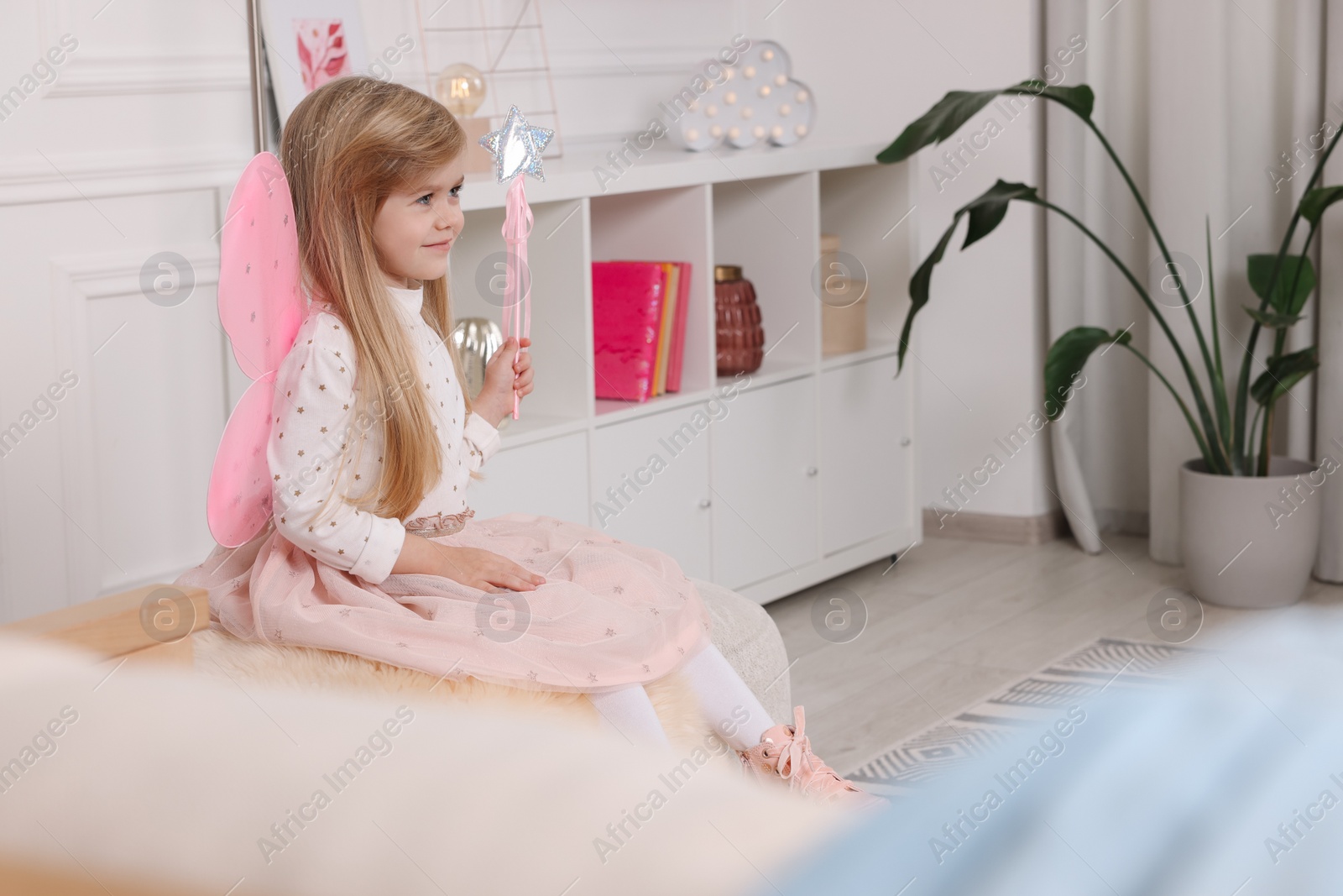 Photo of Cute little girl in fairy costume with pink wings and magic wand at home, space for text