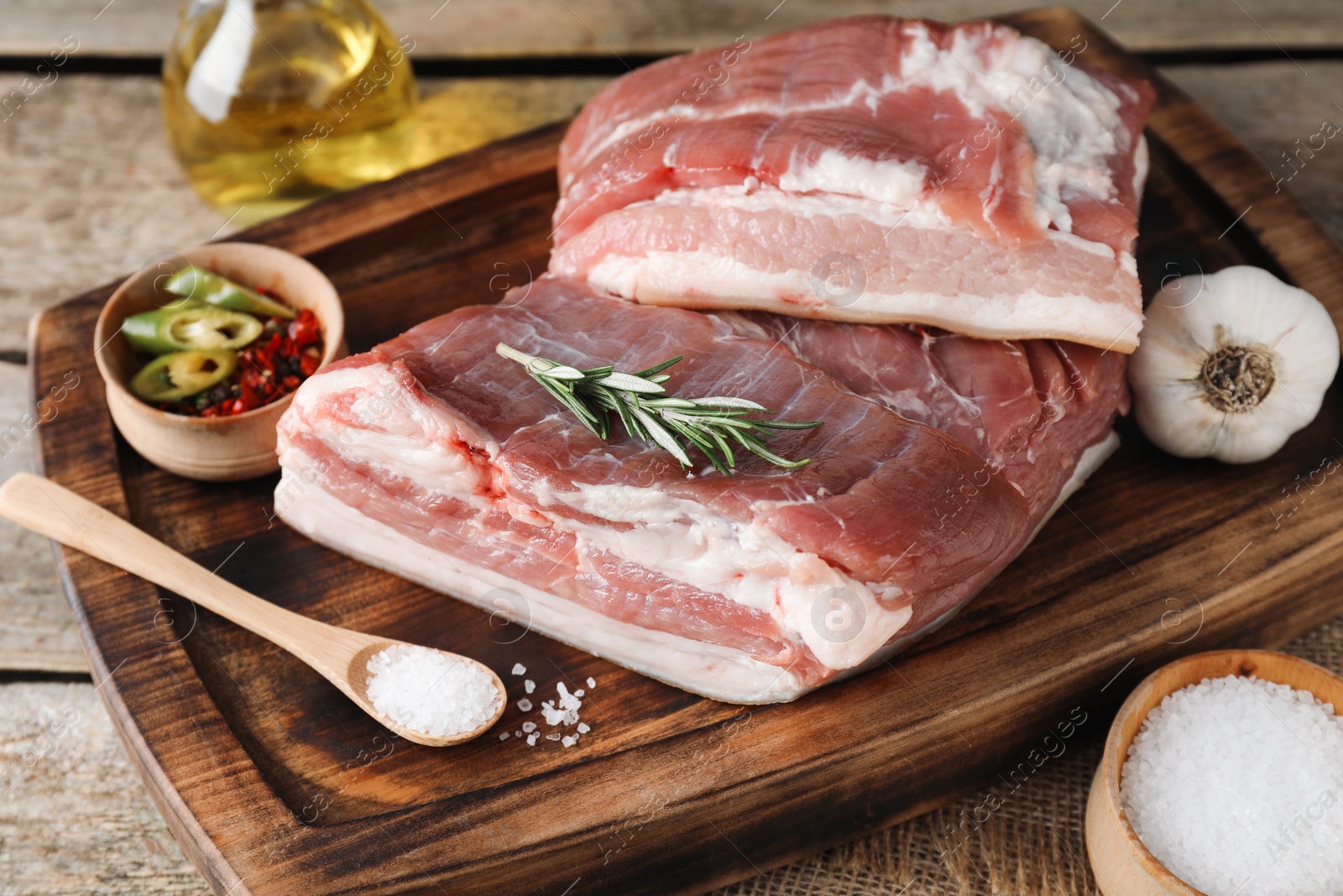 Photo of Pieces of raw pork belly, salt, oil, garlic and rosemary on wooden table, closeup