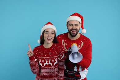 Happy young couple in Christmas sweaters and Santa hats with megaphone on light blue background