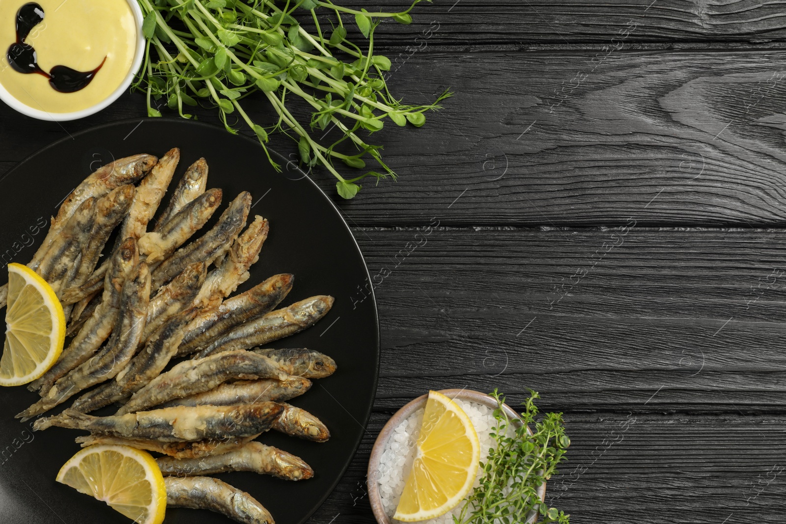 Photo of Delicious fried anchovies with slices of lemon and sauce served on black table, flat lay. Space for text