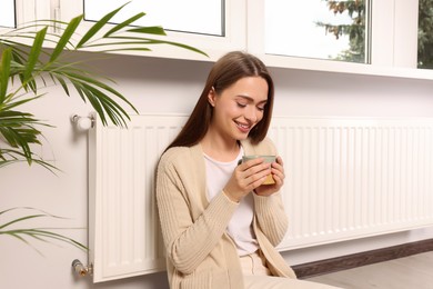 Woman holding cup with hot drink near heating radiator indoors