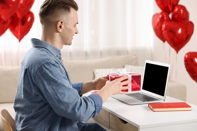 Photo of Valentine's day celebration in long distance relationship. Man holding gift box while having video chat with his girlfriend via laptop at home