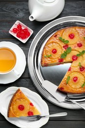 Photo of Delicious cut pineapple pie with cherry and tea served on black wooden table, flat lay