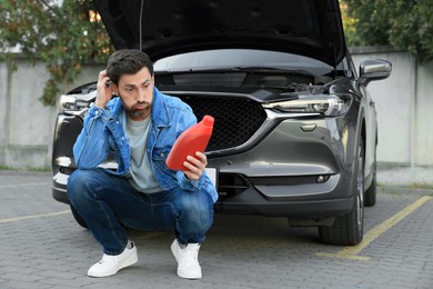 Photo of Puzzled man holding red container of motor oil near car outdoors