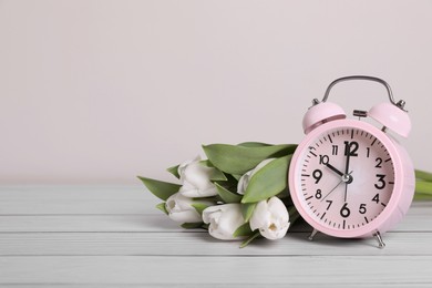 Pink alarm clock and beautiful tulips on white wooden table against light background, space for text. Spring time