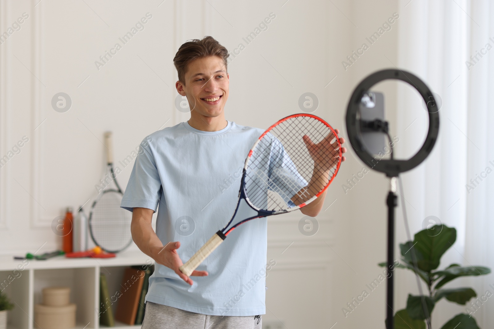 Photo of Smiling sports blogger holding tennis racket while streaming online fitness lesson at home