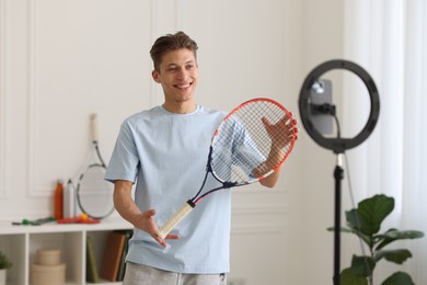 Photo of Smiling sports blogger holding tennis racket while streaming online fitness lesson at home