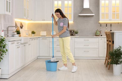 Photo of Happy woman dipping mop into bucket in kitchen