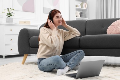 Photo of Happy woman with headphones and laptop on rug in living room