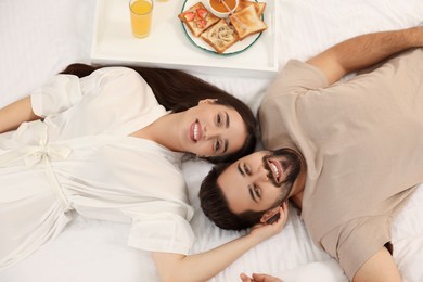 Happy couple lying on bed near white tray with breakfast, above view