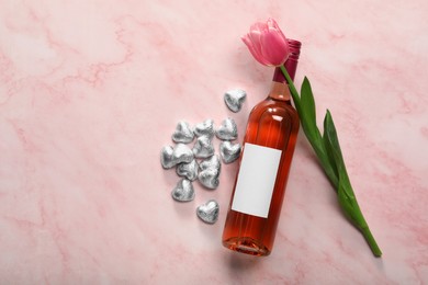 Delicious heart shaped chocolate candies, beautiful tulip and bottle of wine on pink table, flat lay. Space for text
