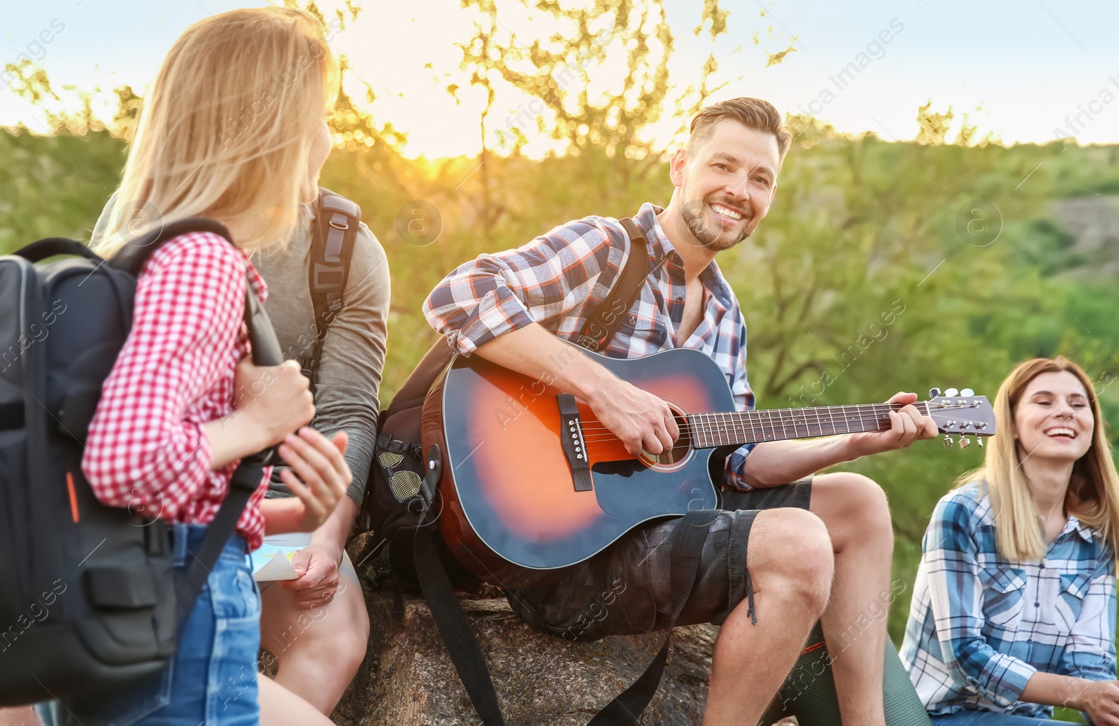 Photo of Young man with backpack playing guitar for his friends in wilderness. Camping season