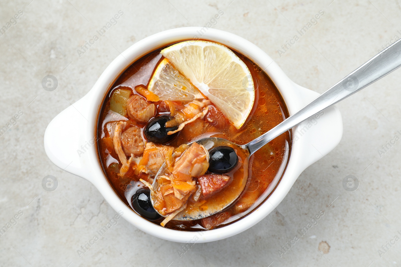 Photo of Meat solyanka soup with sausages, olives, vegetables and spoon in bowl on white textured table, top view