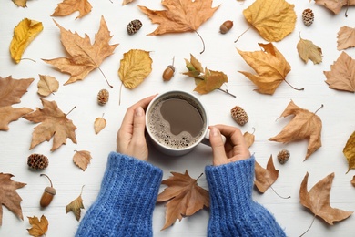 Photo of Woman with cup of hot drink at white wooden table, top view. Cozy autumn atmosphere