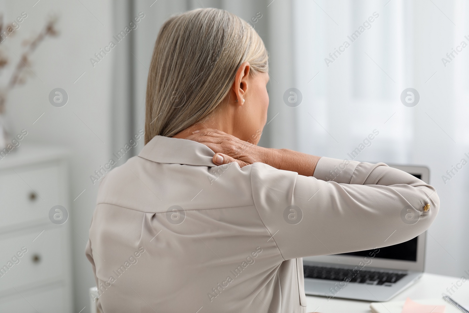 Photo of Woman suffering from neck pain at workplace in room