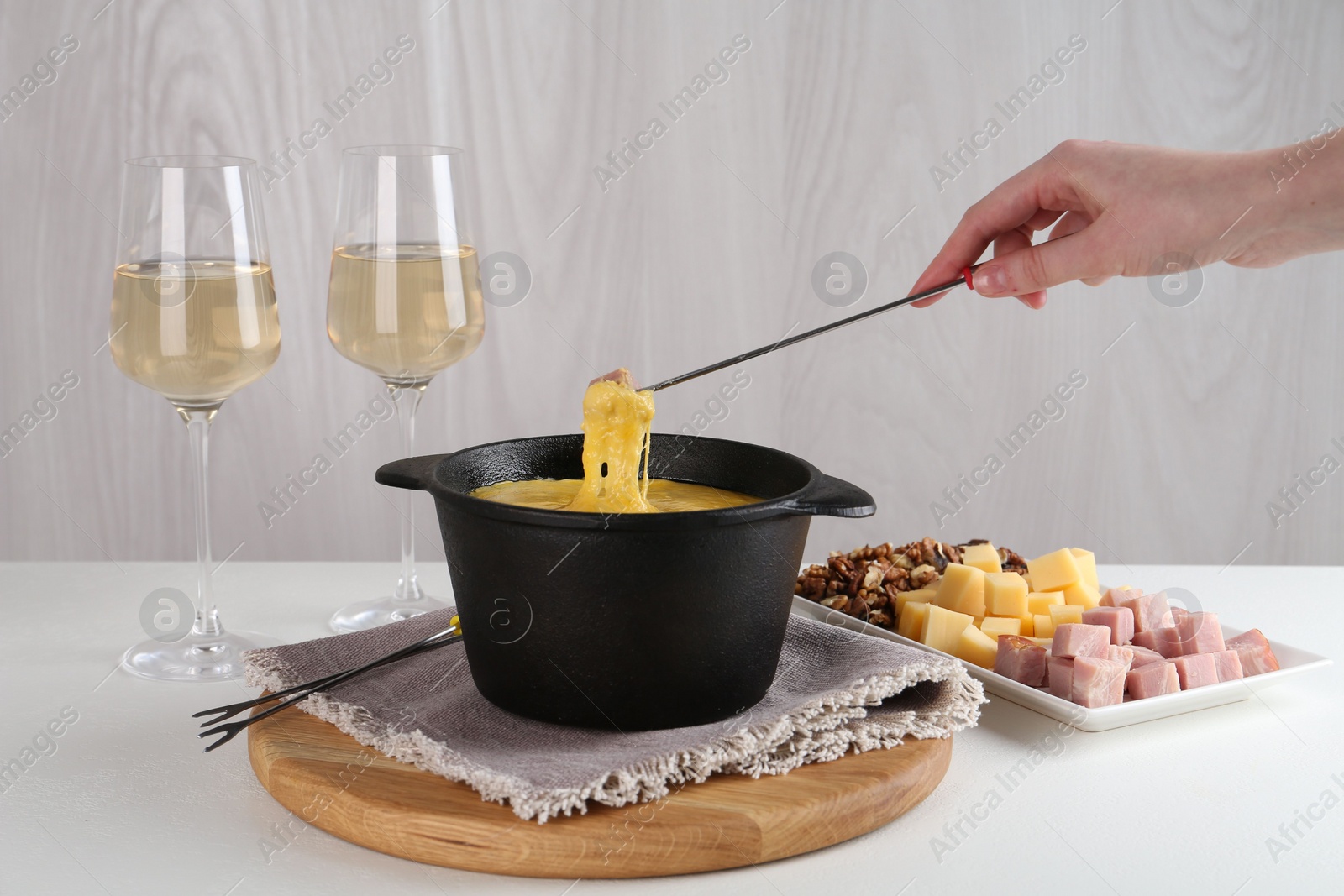 Photo of Dipping piece of ham into fondue pot with tasty melted cheese at white table, closeup