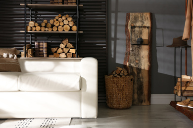 Photo of Shelving unit with stacked firewood and comfortable sofa in stylish room interior