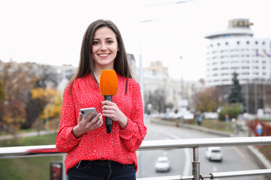 Photo of Young female journalist with microphone and smartphone working on city street. Space for text