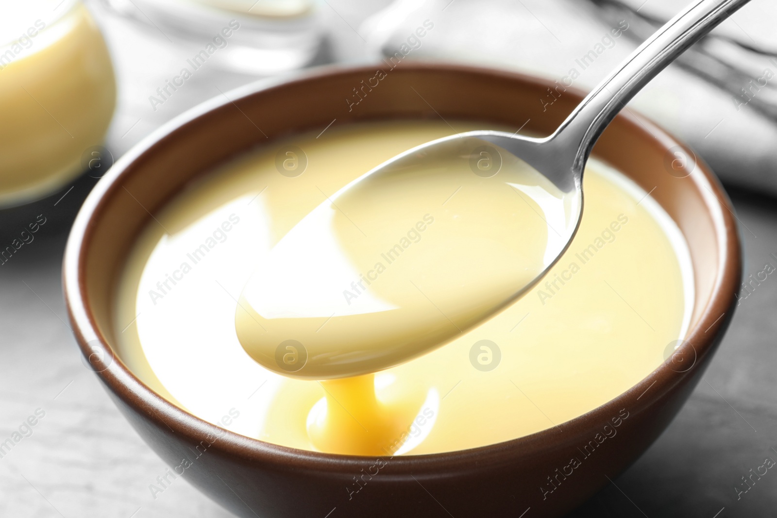 Photo of Spoon of pouring condensed milk over bowl on table, closeup. Dairy products